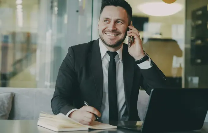 Smiling man in suit talking on a phone while taking notes at his laptop in a bright modern office featured on Town Service Zone